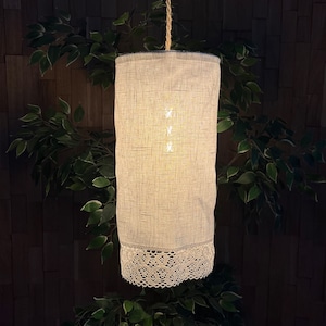 May include: A white linen lampshade with a lace trim hangs from a cord. The lampshade is illuminated from within, casting a warm glow.
