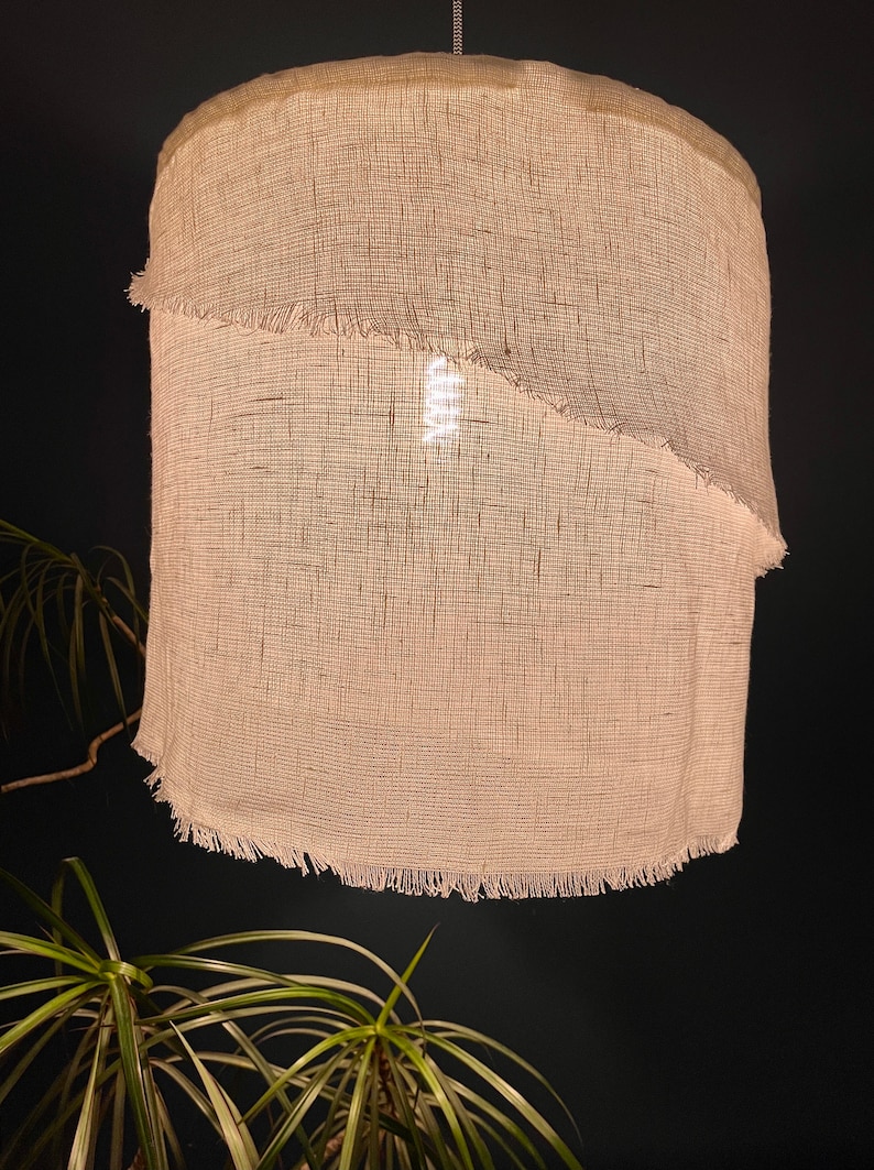 Pu&ograve; includere: Un paralume bianco cilindrico in tessuto tessuto con un bordo sfrangiato. Il paralume &egrave; illuminato dall'interno, proiettando una luce calda.