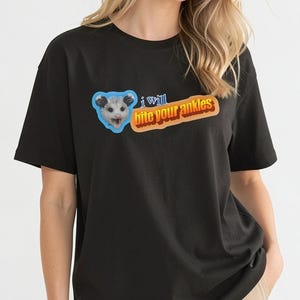 Peut inclure: Un t-shirt noir avec un motif représentant une tête d'opossum dans une forme bleue à côté d'une bulle orange contenant le texte "i will bite your ankles" dans une police rétro en gras avec un effet d'ombre portée sur la poitrine.