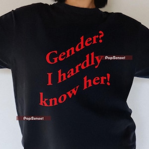 May include: Black t-shirt with the red text "Gender? I hardly know her!" The shirt is a dark colour and has a casual style. The text is in a bold, slightly curved font. The shirt is worn by a person.