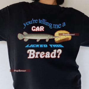 May include: Black t-shirt with a graphic of a fish licking a piece of bread. The text on the shirt reads "You're telling me a GAR licked this Bread?"