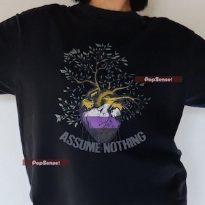 May include: Black t-shirt featuring a graphic of a tree growing from a heart shape, with the text "ASSUME NOTHING" in a stylized font. The design incorporates shades of yellow, purple, and green.