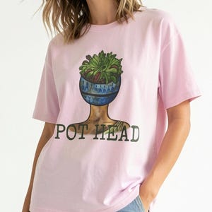 Może przedstawiać: Jasnoróżowy t-shirt z krótkim rękawem z nadrukiem graficznym przedstawiającym niebieską, wzorzystą doniczkę z zielonymi sukulentami umieszczoną na szyi i ramionach. Napis POT HEAD jest wydrukowany ciemnozielonymi wielkimi literami pod grafiką.