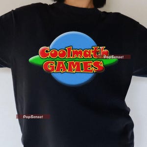以下が含まれることがあります： 「Coolmath Games」と書いてある青、緑、赤のロゴがプリントされた黒いTシャツ。