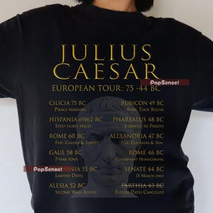 May include: Black t-shirt featuring the text "JULIUS CAESAR EUROPEAN TOUR: 75-44 BC" in gold lettering. The shirt also includes a bust of Julius Caesar and a list of locations and dates from his tour.