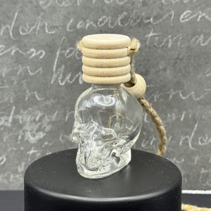 Może przedstawiać: Przezroczysta szklana butelka w kształcie czaszki z drewnianym korkiem i sznurkiem z naturalnego włókna. Butelka stoi na czarnej cylindrycznej podstawie. Tło to szara powierzchnia z białym pismem.