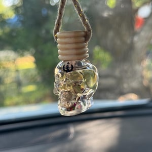 Könnte beinhalten: Ein durchsichtiger, totenkopfförmiger Auto-Lufterfrischer aus Glas, gefüllt mit einer hellgelben Flüssigkeit. Die Flasche zeigt ein kleines schwarzes Symbol der Rebellenallianz aus Star Wars auf der Stirn und hat einen gerippten Holzverschluss.