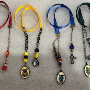 May include: Four handmade ribbon bookmarks with decorative charms. The ribbons are red, yellow, blue, and green. Each bookmark has a different charm, including a lightning bolt, a badger, a key, and a snake.