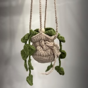 May include: A handmade, hanging crochet plant holder. The beige-colored pot is suspended by four beige cords. Green crochet leaves cascade from the top and sides of the pot, creating a decorative, natural look. A wooden bead is visible at the top.