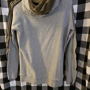 May include: A grey hoodie with an olive green hood lining. The sleeves have a decorative stripe in shades of brown and green. The hoodie is hanging on a white hanger against a black and white checked background.