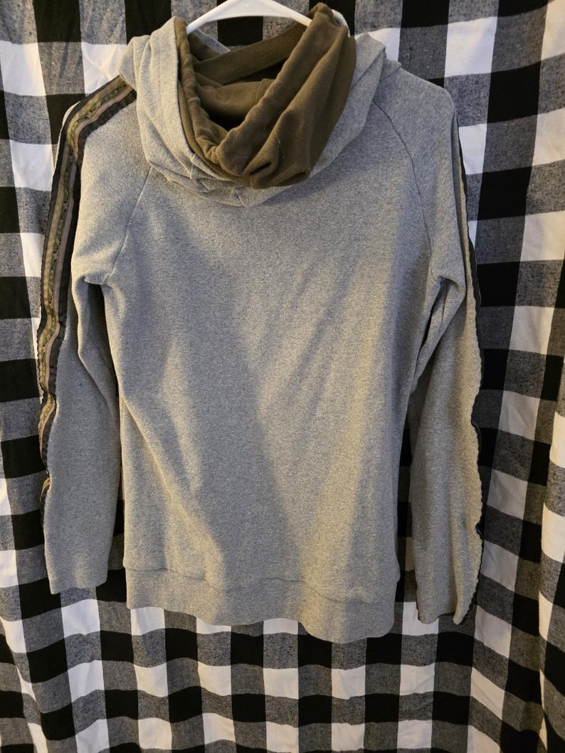 May include: A grey hoodie with an olive green hood lining. The sleeves have a decorative stripe in shades of brown and green. The hoodie is hanging on a white hanger against a black and white checked background.