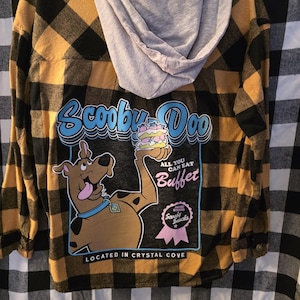 Puede incluir: Una camisa de manga larga con capucha y un estampado de cuadros amarillos y negros. La parte trasera presenta un gráfico de Scooby-Doo con el texto "Scooby Doo All You Can Eat Buffet" y "Located in Crystal Cove".