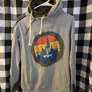 May include: Grey hoodie featuring a Disneyland Resort graphic. The graphic is a circular design with a castle silhouette and the text "Happiest Place on Earth". The hoodie has a drawstring hood and a zip detail.