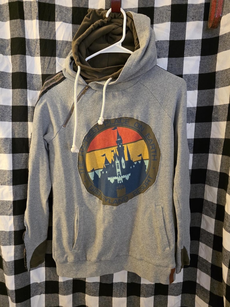 May include: Grey hoodie featuring a Disneyland Resort graphic. The graphic is a circular design with a castle silhouette and the text "Happiest Place on Earth". The hoodie has a drawstring hood and a zip detail.