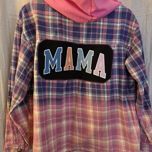 Puede incluir: Una camisa de franela a cuadros con capucha rosa. La camisa presenta un diseño descolorido de azul a rosa. Un parche negro en la espalda muestra la palabra "MAMA" en letras blancas, azules y rosas.
