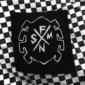 Puede incluir: Parche blanco y negro con un diseño de telaraña estilizado y las letras "SFMN" en el centro.