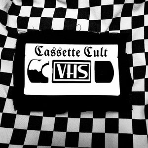 May include: Black and white cassette tape patch with the text "Cassette Cult" and "VHS" in a vintage style font.