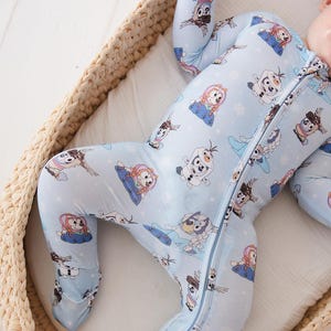 May include: Light blue baby sleeper featuring a repeating pattern of cartoon characters. The sleeper has a full-length zipper and covers the feet. The baby is lying in a woven basket with a white blanket.
