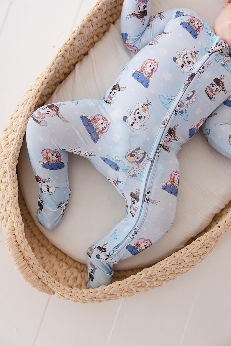 May include: Light blue baby sleeper featuring a repeating pattern of cartoon characters. The sleeper has a full-length zipper and covers the feet. The baby is lying in a woven basket with a white blanket.