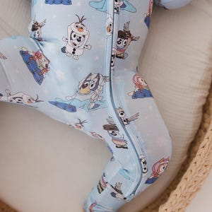 May include: Light blue baby sleeper featuring a repeating pattern of cartoon characters and snowflakes. The one-piece garment has a full-length zipper and long sleeves. The sleeper is made of a soft material and is designed for comfort.