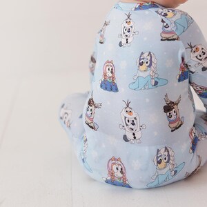 May include: Light blue baby one-piece pajama with a repeating pattern of cartoon characters, including a snowman and a dog with a blue dress and a braid. The pajama has long sleeves and legs. The background is white.