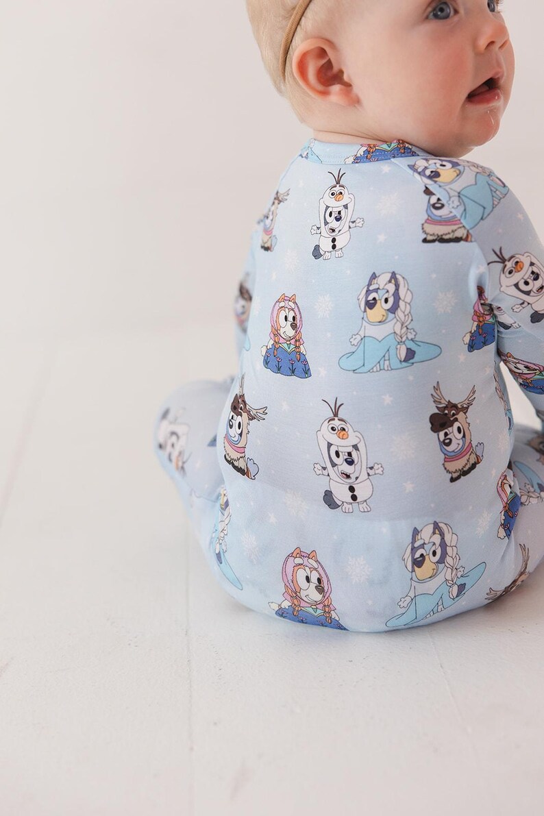 May include: Light blue baby one-piece pajama with a repeating pattern of cartoon characters, including a snowman and a dog with a blue dress and a braid. The pajama has long sleeves and legs. The background is white.
