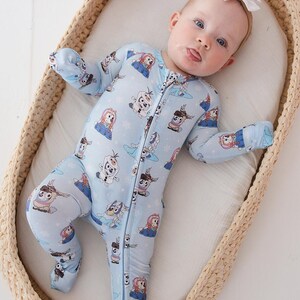 May include: A baby wearing a light blue footed onesie with a zipper closure. The onesie features a repeating pattern of cartoon characters. The baby is lying in a woven basket and wearing a white bow.