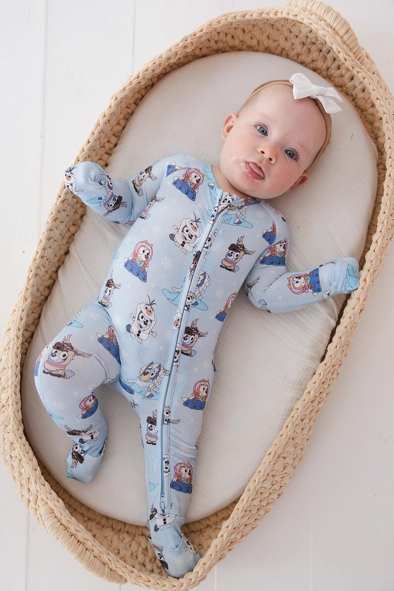May include: A baby wearing a light blue footed onesie with a zipper closure. The onesie features a repeating pattern of cartoon characters. The baby is lying in a woven basket and wearing a white bow.