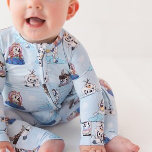 May include: A light blue baby sleeper featuring a zipper closure and long sleeves. The sleeper is adorned with a repeating pattern of cartoon characters and snowflakes. The baby is wearing a light brown headband.