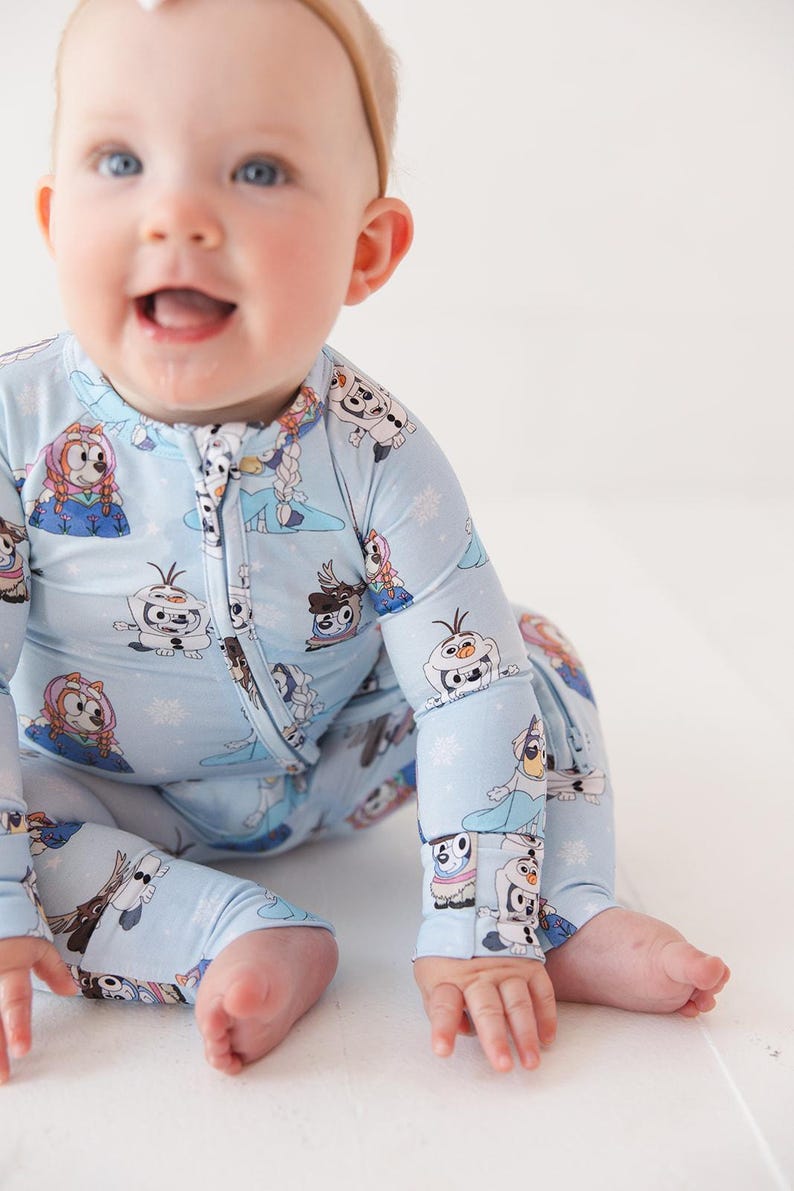 May include: A light blue baby sleeper featuring a zipper closure and long sleeves. The sleeper is adorned with a repeating pattern of cartoon characters and snowflakes. The baby is wearing a light brown headband.
