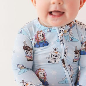 May include: Light blue baby one-piece pajama with a repeating pattern of cartoon characters and snowflakes. The pajama has a full-length zipper and long sleeves. The baby is smiling.