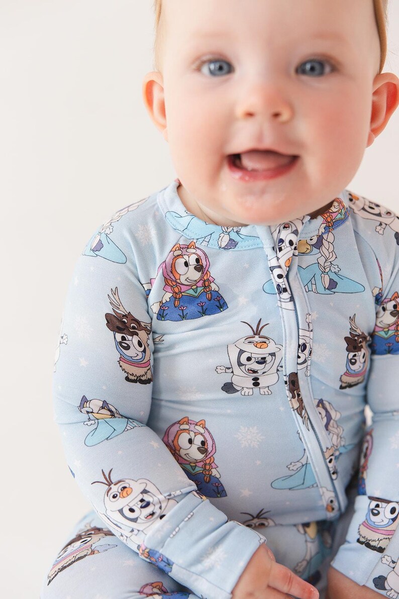 May include: Light blue baby one-piece pajama with a repeating pattern of cartoon characters and snowflakes. The pajama has a full-length zipper and long sleeves. The baby is smiling.