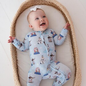 May include: A baby wearing a light blue onesie with a zipper and a pattern of cartoon characters. The baby is lying in a woven basket with a white interior and wearing a white bow headband. The baby is smiling.