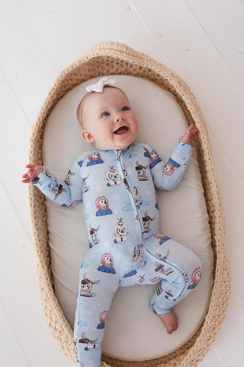 May include: A baby wearing a light blue onesie with a zipper and a pattern of cartoon characters. The baby is lying in a woven basket with a white interior and wearing a white bow headband. The baby is smiling.