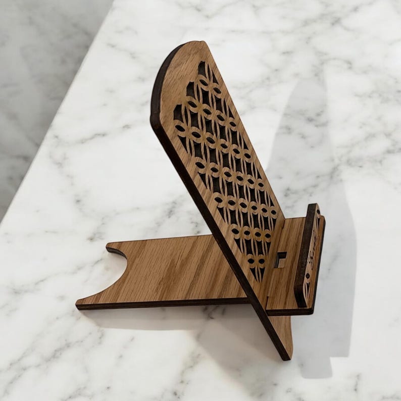 May include: Wooden phone stand with a decorative laser cut design. The stand is made of light brown wood and has a small cutout for a phone.