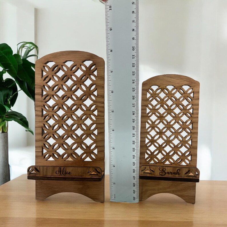 May include: Two wooden phone stands with intricate laser cut designs. The stands are personalized with the names "Alice" and "Sarah".