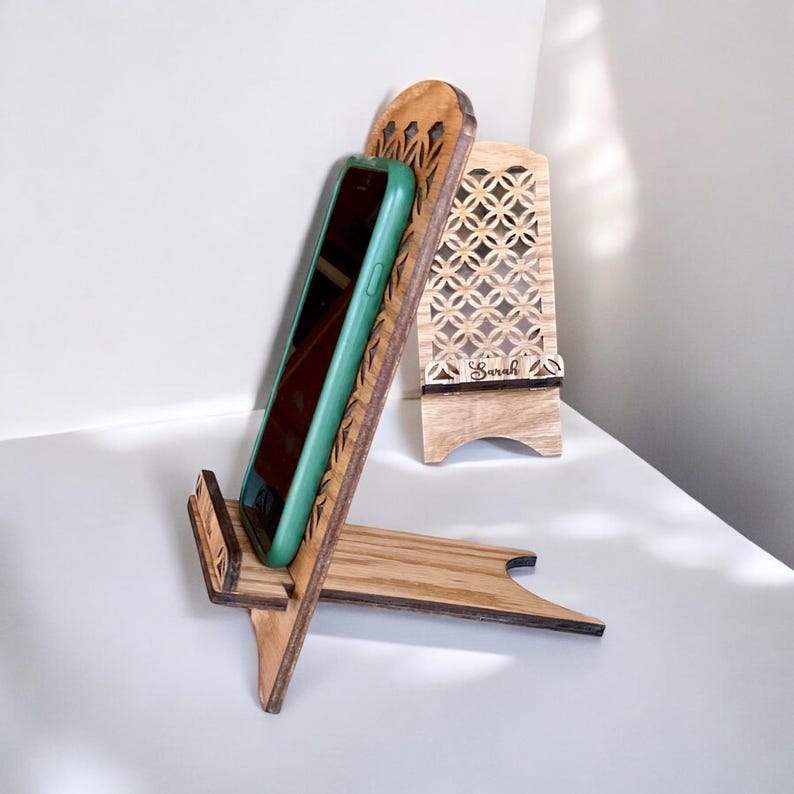 May include: A wooden phone stand with a decorative cutout design. The stand is made of light brown wood and has a small, rectangular base. A teal phone is propped up in the stand. The stand is engraved with the name "Sarah".