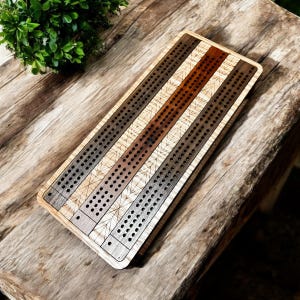 May include: A wooden cribbage board with a striped design. The board features alternating stripes of light, medium, and dark brown wood, with rows of drilled holes for pegs. The board is rectangular with rounded corners.