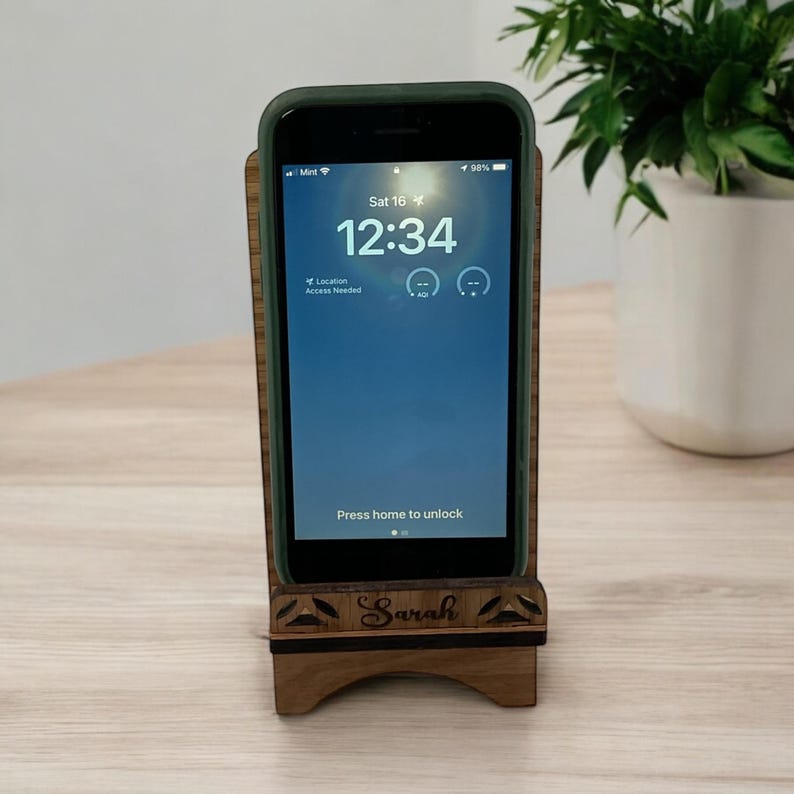 May include: A wooden phone stand with a personalized name "Sarah" engraved on the front. The stand is holding a smartphone with a lock screen displaying the time 12:34.