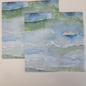 May include: Two square paper napkins with a watercolor design. The napkins feature a landscape of blue, green, and white hues, resembling clouds and mountains. The napkins are placed on a white surface.