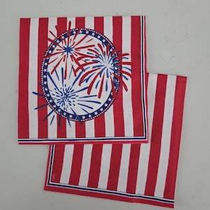 May include: Two square paper napkins featuring a red and white vertical striped pattern. One napkin displays a central circular emblem with a blue border containing white stars, enclosing a graphic of red and blue fireworks bursting outward.