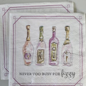 May include: Two white paper napkins with a watercolor illustration of four champagne bottles in shades of pink, gold, and green. The napkins have a purple geometric border and the text "NEVER TOO BUSY FOR fizzy".