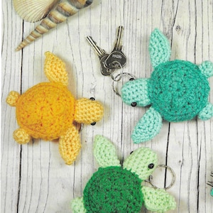 May include: Three handmade crochet turtle keyrings in yellow, teal, and green. Each turtle has a round body, flipper-like appendages, and a metal keyring. The keyrings are displayed on a light wood-grain surface.