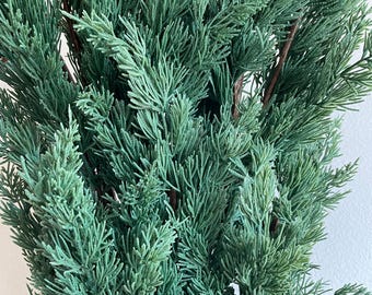 May include: A bunch of artificial cedar sprigs in a deep green color. The sprigs are arranged in a cluster, creating a full and lush appearance.