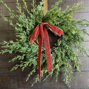 May include: A fresh evergreen wreath with vibrant green foliage and a deep red velvet ribbon. The wreath is a circular shape, perfect for seasonal decor. The ribbon is tied in a bow and hangs down.