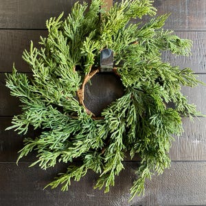 May include: A green cedar wreath with a brown woven base, hanging on a dark brown wooden door.