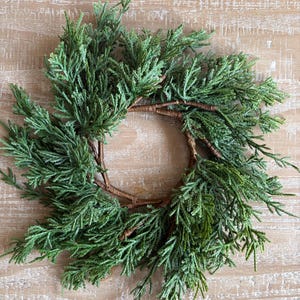 May include: A small circular wreath constructed from a brown twig frame adorned with dense, textured green cedar foliage. The evergreen branches are arranged in a radial pattern, creating a full, layered appearance around the central opening.