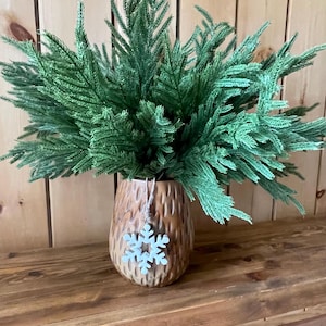 May include: A decorative arrangement featuring a textured brown vase filled with green faux evergreen branches. A snowflake ornament hangs from the vase. The arrangement sits on a wooden surface against a wooden wall.