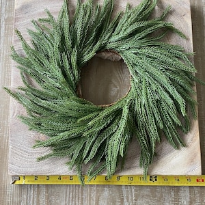 May include: A green, leafy wreath with a natural, circular design. The wreath is approximately 16 inches in diameter and is placed on a wooden square. A yellow measuring tape is placed across the bottom of the wooden square.