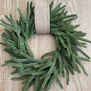 May include: A circular green wreath made of faux evergreen branches. A wide, tan burlap ribbon is used to hang the wreath. The wreath is displayed against a light-colored, wood-grain background.